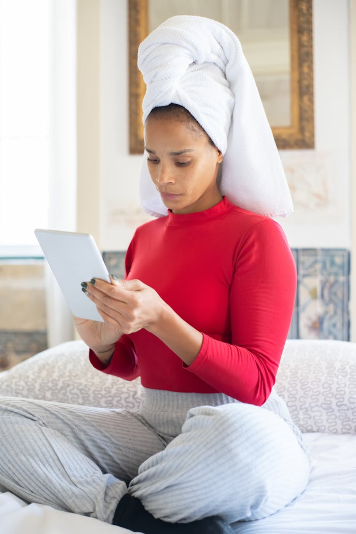 African American woman with head towel sitting on bed, reading a tablet device indoors.